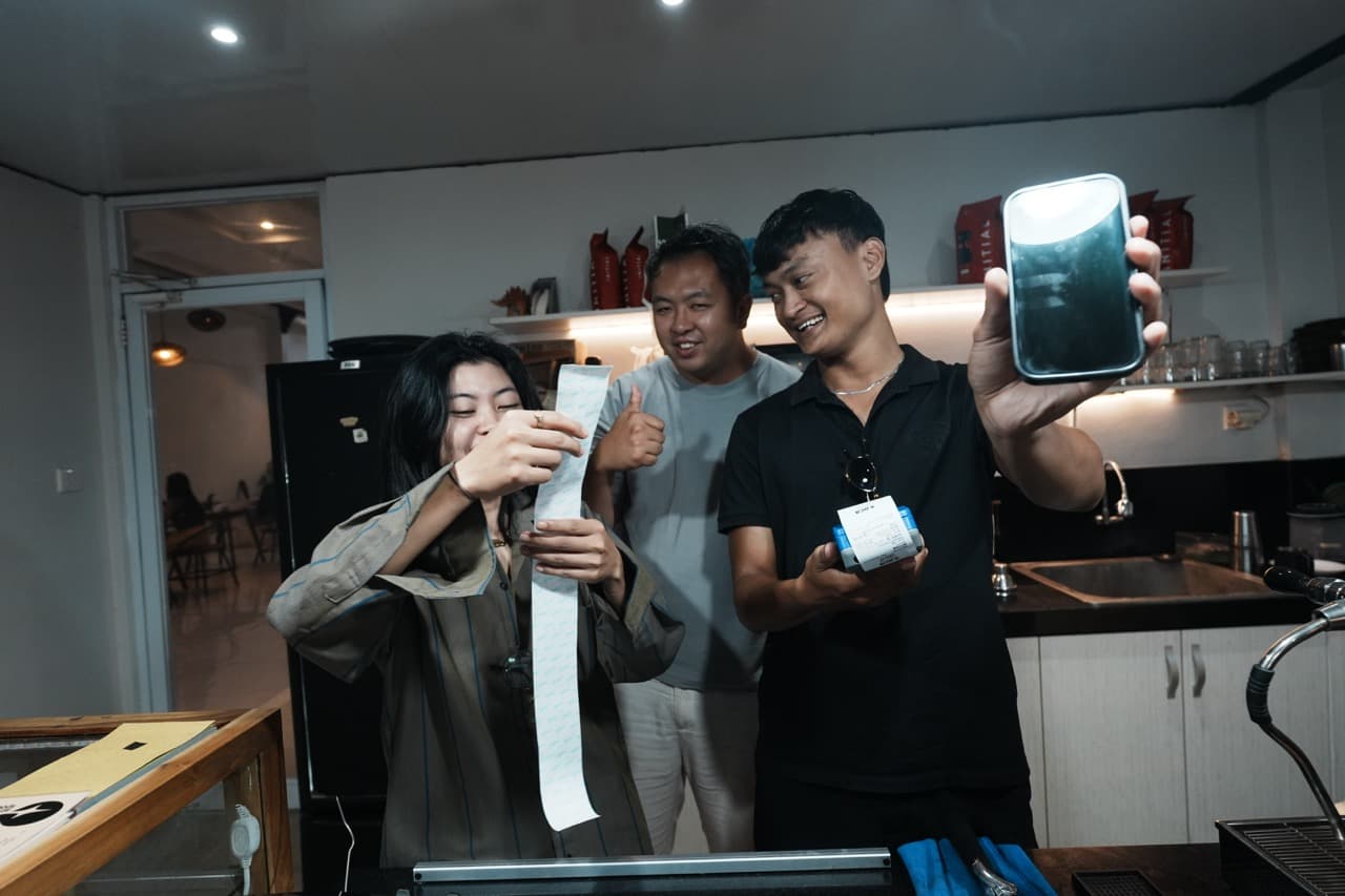 Trois membres d'une équipe de café rient derrière le comptoir : l'une tient un long ticket de caisse, un autre montre un téléphone et une petite imprimante de tickets.